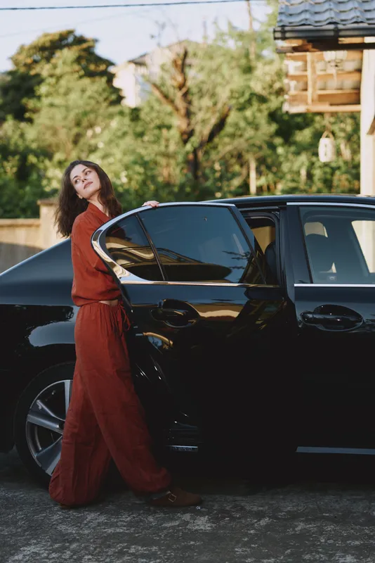 Elegant young woman in a flowing red dress stands beside a sleek black car outdoors, looking back over her shoulder with a soft, confident smile under bright daylight