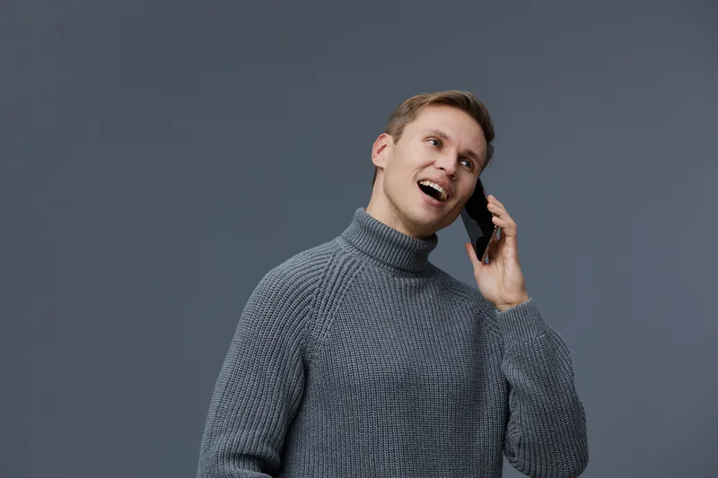 Smiling man talking on a mobile phone, wearing a gray sweater, against a plain dark background, studio portrait, casual style, happiness, communication, technology, facial expression.