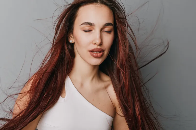 Young woman with long red hair and closed eyes posing calmly in white sleeveless top on grey background. Beauty portrait with natural makeup and serene expression in studio lighting.