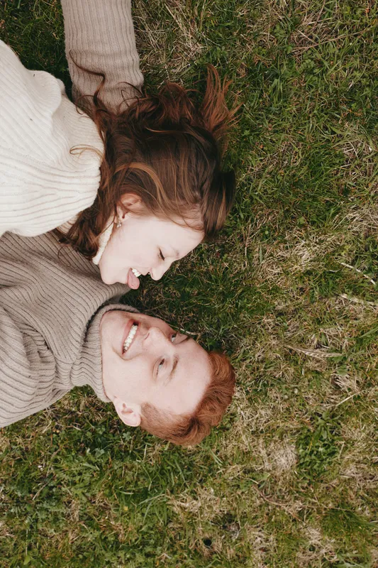 Cheerful couple lying on lush grass outdoors, faces close, sunlit moment, cozy sweaters, relaxed mood, natural colors, candid lifestyle scene, affectionate connection, together
