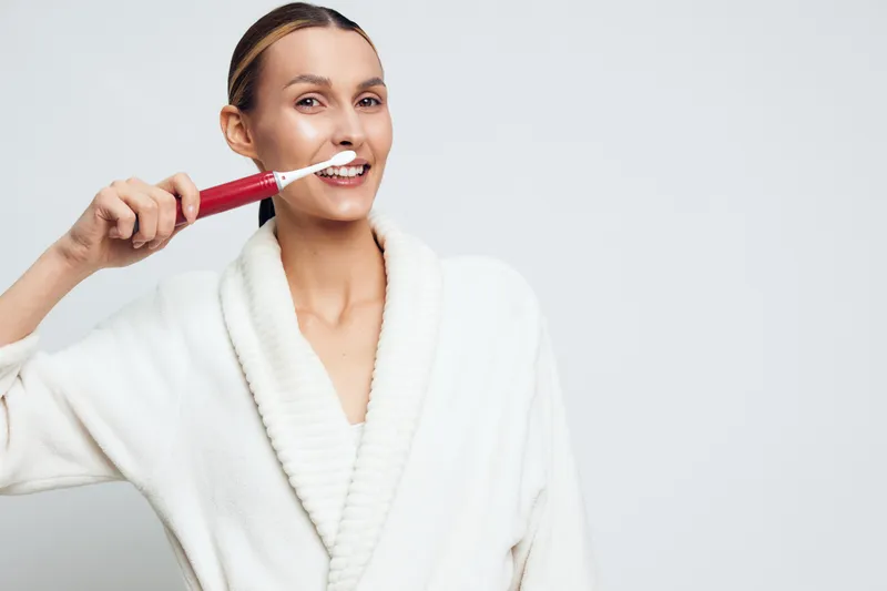 Young woman in white bathrobe smiling while brushing teeth with electric toothbrush, clean background, personal hygiene, morning routine, dental care concept.