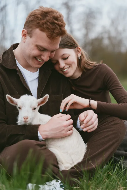 A couple outdoors gently cuddle a white goat kid in a sunlit field, smiling warmly, showcasing a rustic lifestyle and bond with animals.