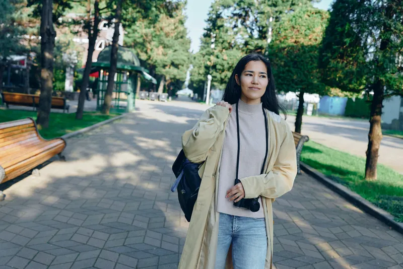 Woman in light coat with camera walks through a sunny park, casual street style, greenery, and relaxed mood in an urban outdoor scene featuring bright light, breeze, and casual fashion vibe for