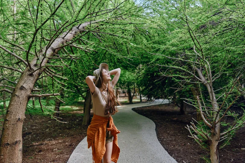 Woman walking on winding path under green trees in a forest park, relaxed and carefree with long hair and boho dress, enjoying nature and sunlight through branches.