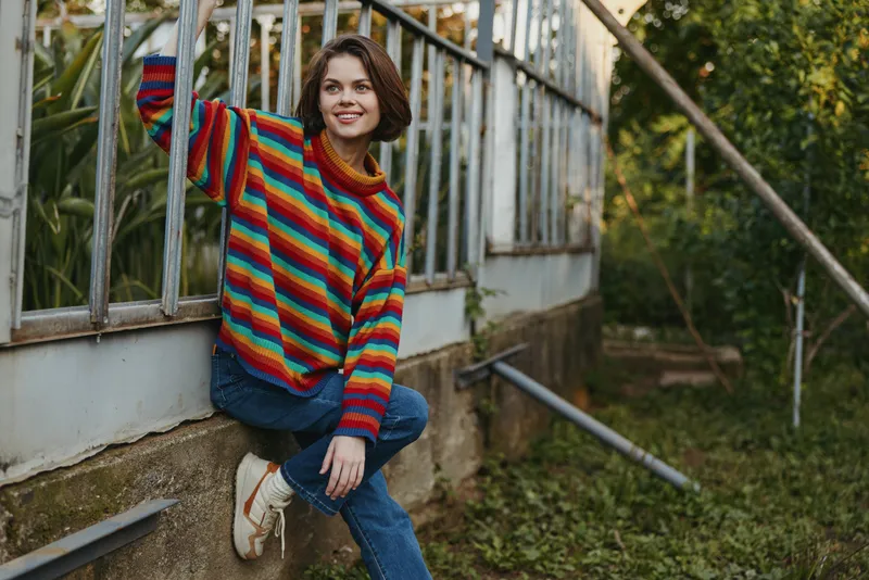 Woman sweater greenhouse smile casual outdoors jeans sitting on ledge, young female model wearing colorful striped jumper and sneakers, relaxed pose by glass wall in garden setting.