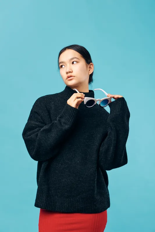 Young woman in black sweater holding white sunglasses and looking sideways on blue background. Female model wearing red skirt with casual fashion style and natural expression.