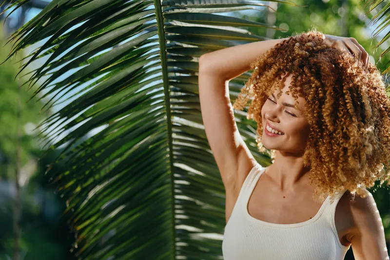 Smiling woman with curly hair enjoys sunny tropical outdoors wearing white tank top near green palm leaves, expressing happiness and relaxed summer vibe.