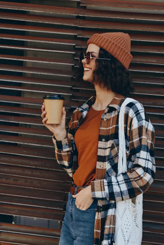 Woman holding coffee cup with smile in urban street, wearing beanie, sunglasses and plaid shirt with tote bag, candid lifestyle capture with authenticity, golden hour glow and mindful living.