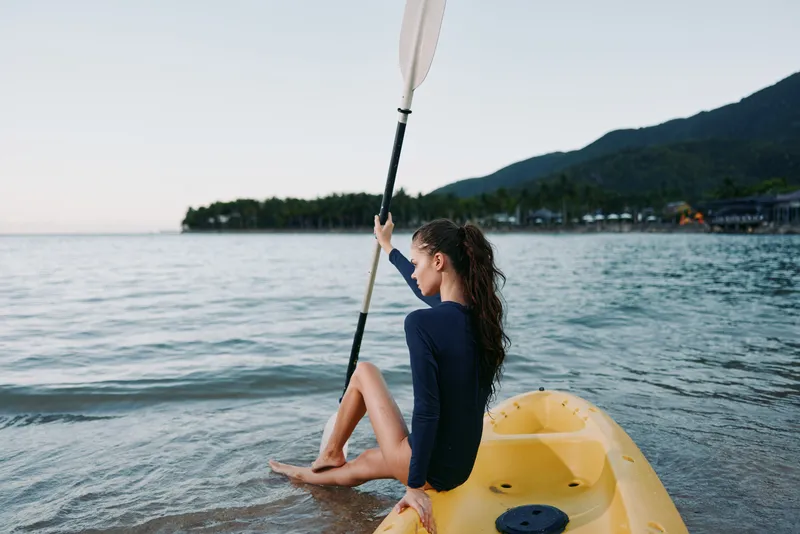A young woman sits on a bright yellow kayak near the shore, raising a paddle as calm water laps the hull during a serene sunset session.