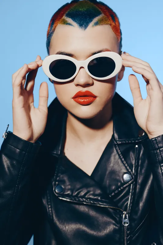 Woman with colorful short hair and white sunglasses wearing a black leather jacket, bright red lipstick, and an edgy stylish look on a blue background.