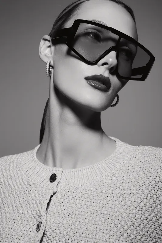 Stylish young white woman in large geometric sunglasses and textured sweater poses in black and white studio portrait. Concept of fashion, elegance, and modern accessories.
