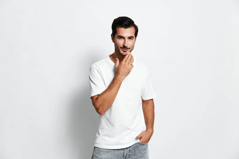 Handsome thoughtful young man in white t-shirt looks at the camera with a slight smile, standing against a plain solid background. Casual lifestyle concept.