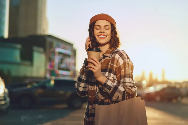Woman holding coffee and shopping bag, smile in urban street at golden hour wearing beanie; candid authenticity and candid lifestyle mood with golden hour glow, mindful living, emotional storytelling.