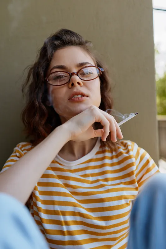 Young woman with round glasses sits at home wearing a yellow striped shirt, resting her chin on her hand with a cigarette, presenting a casual candid mood in a relaxed indoor setting