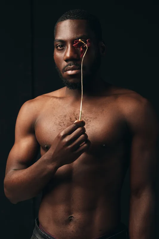 African man with dark skin and muscular body holding dried flower in creative conceptual visual style. Portrait with artistic expression and minimalistic dark background for emotional impact.