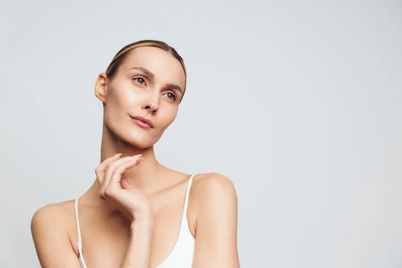Woman skincare beauty wellness self care close up face portrait touching cheek on light gray background soft studio lighting calm relaxing mood with bare shoulders minimal composition