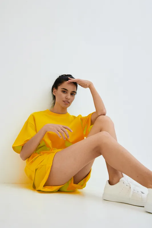 Young woman wearing bright yellow casual clothes and white sneakers sits relaxed against white minimalist background, showcasing modern sporty style and calm expression.