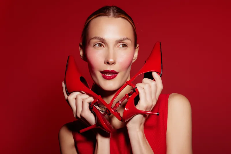 Young woman in red dress holds red high heels near face, posing on red background. Concept of fashion, style, elegance, and vibrant color contrast in studio portrait.