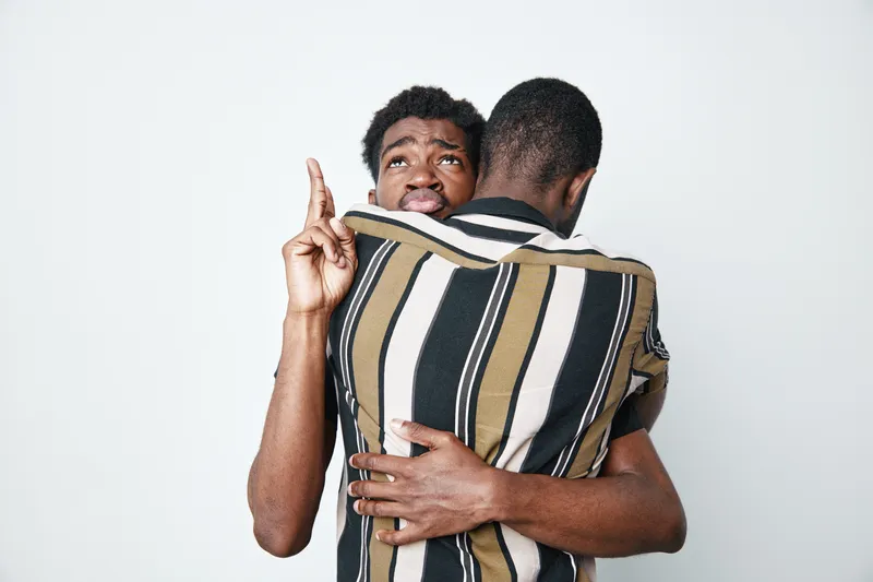 Two african men with dark skin hugging each other, one looking worried and pointing finger, emotional support and connection in studio with white background and casual clothing.