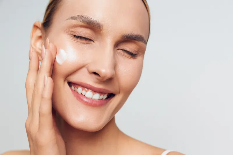 A young adult brunette woman applies moisturizer to her clean, radiant skin, smiling softly as she performs a simple skincare routine in a bright studio setting.