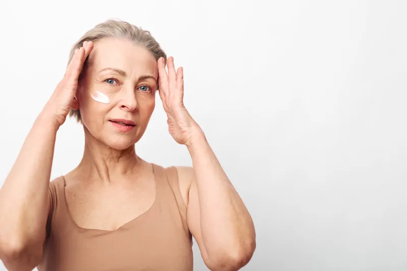 Mature woman with grey hair applies white face cream on cheeks, wearing beige tank top. Isolated on white background. Concept of skincare, aging skin care, beauty routine and self-care.