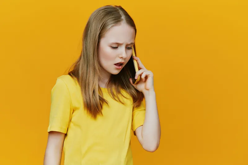 Concerned girl in a yellow shirt talking on the phone against a bright yellow background, capturing emotions of surprise and worry, ideal for communications concepts