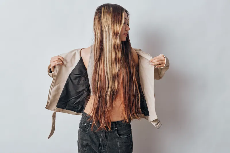 Young woman with long hair removing beige jacket and wearing sleeveless top standing against white background for fashion and style concept.
