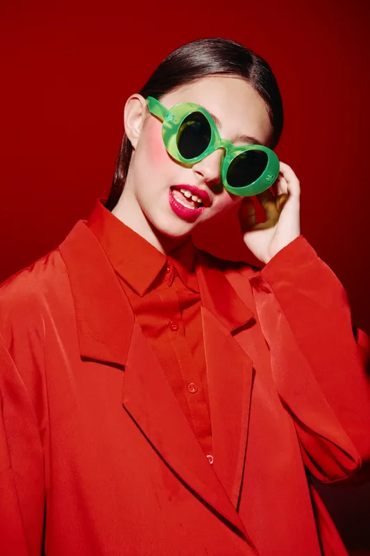 Young woman wearing red oversized blazer and shirt poses confidently with bright green sunglasses on red background. Editorial concept of beauty fashion creativity.