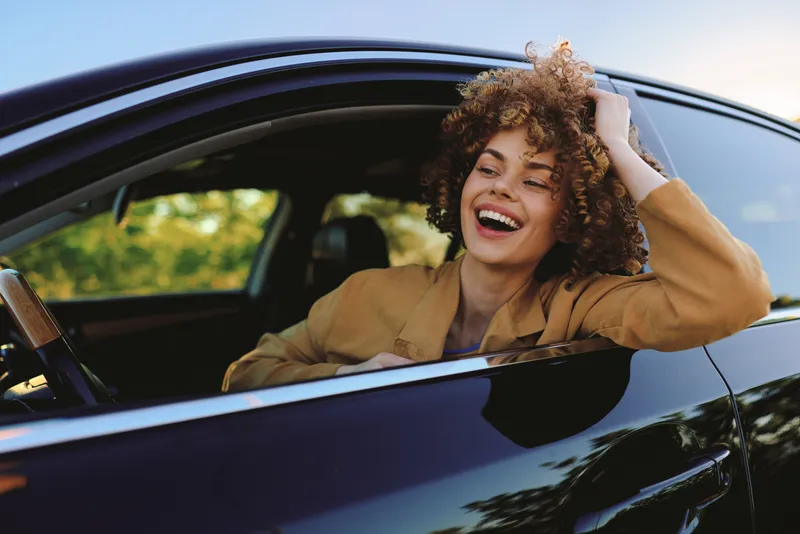 Cheerful woman inside a car, laughing with head tilted back, sunlit moment, casual chic style, retro film color vibe, candid lifestyle shot, warm tones, vibrant outdoors scene in natural light