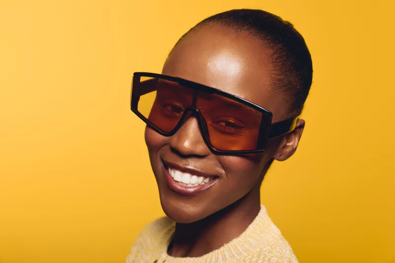Smiling dark skinned African woman with short hair wearing large stylish sunglasses and yellow sweater. Bright yellow background enhances modern fashion and joyful mood concept.