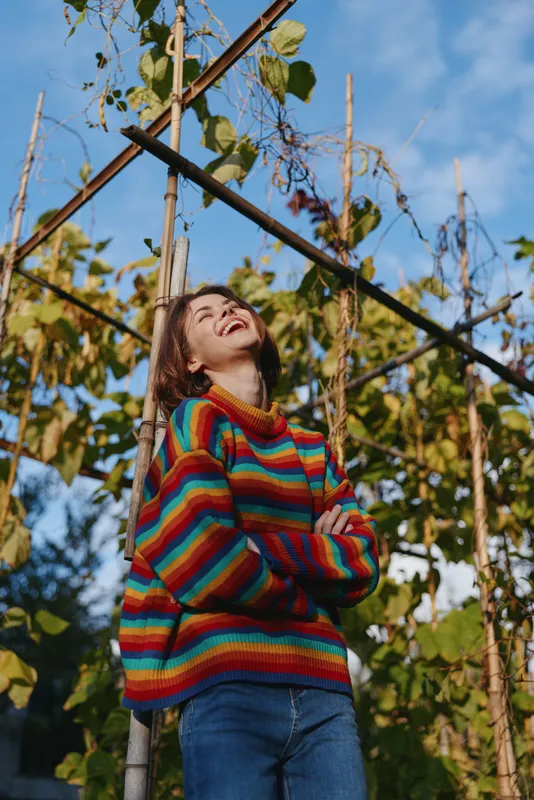 Young adult woman smiling and laughing in a rainbow striped sweater, casual outfit leaning on a garden trellis under blue sky in outdoor lifestyle portrait with warm sunlight.