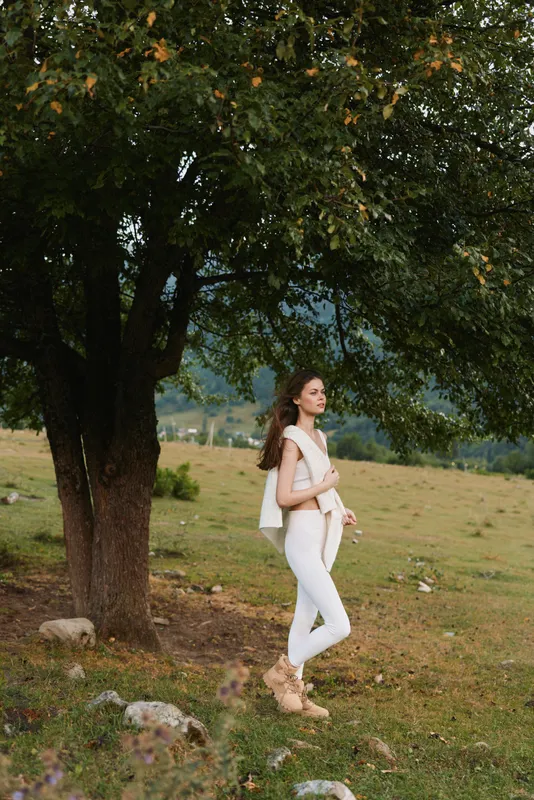 Woman lifestyle outdoors, standing near a tree in a sunlit meadow, wearing white outfit, relaxed pose, gentle breeze, natural colors, peaceful countryside.