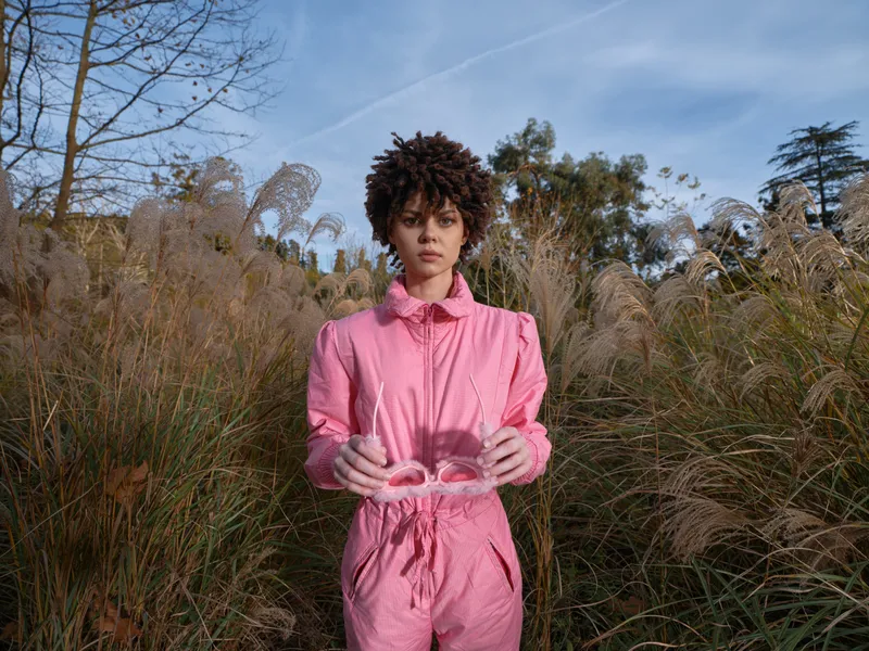 Fashionable woman in a pink suit stands in tall grasses outdoors, holding pink sunglasses with a calm expression under a clear blue sky, expressive portrait of bold style and natural light.