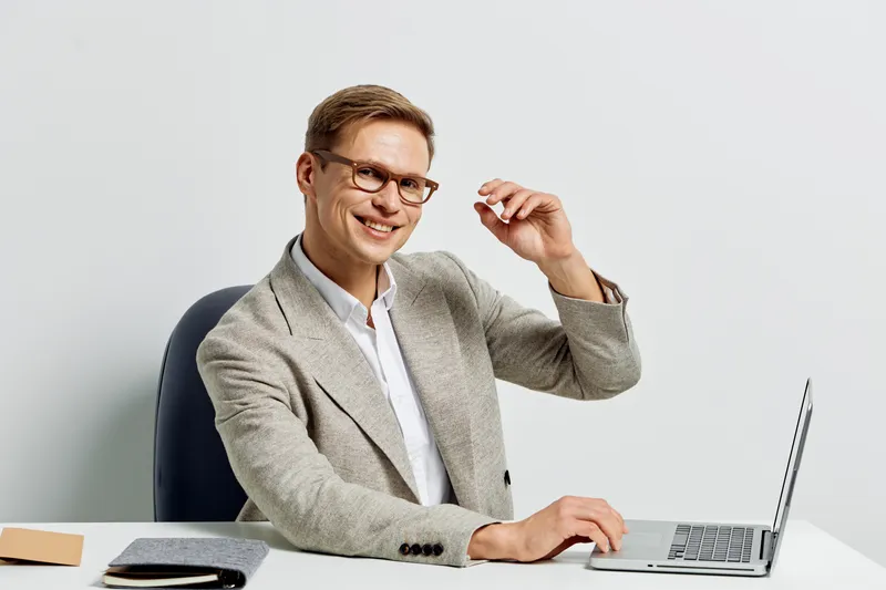 Confident young man in smart casual attire, wearing glasses, smiling and looking at camera while sitting at desk with laptop on plain white background. Business, success, professional concept.