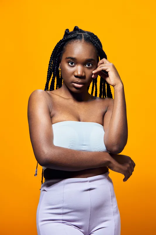 Young African woman with braids poses thoughtfully against a vibrant orange background, wearing a light tube top and pastel pants, expressing confidence and beauty