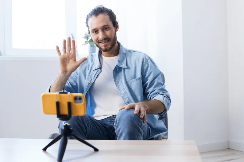 Young man smiling and waving during a video call at home, wearing casual blue clothing, creating a friendly and engaging atmosphere for online communication