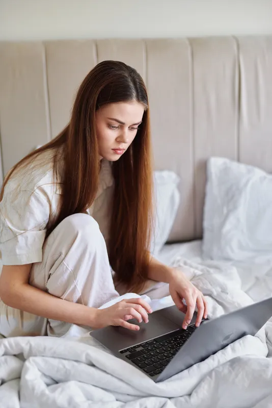 Woman on bed working on laptop in white pajamas at home bedroom, focused young adult typing on computer, remote work or studying, casual morning routine and cozy interior.