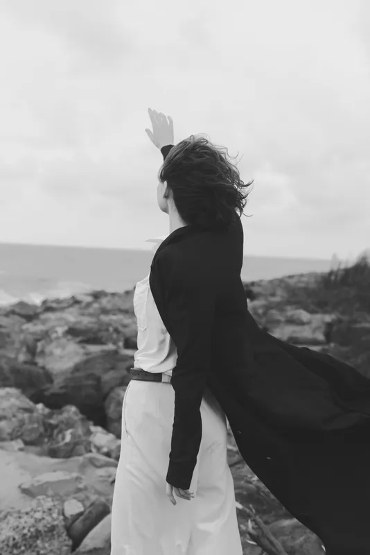 Wind swept woman on a rocky coast in a flowing white dress and dark coat raises arms toward the sea, a bold windblown moment of freedom.