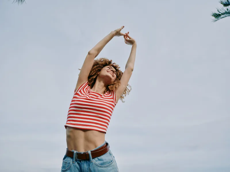 A joyful young woman stretches arms overhead under a bright sky, wearing a striped crop top and jeans, radiating carefree summer energy and confident freedom.