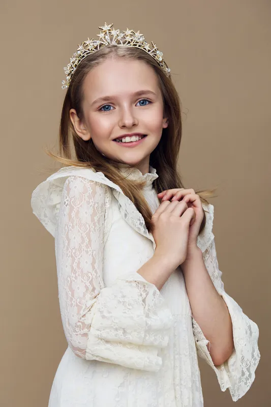 Young girl in elegant lace dress with tiara, smiling against a warm beige background, embodying innocence and joy, perfect for children s fashion or royalty themed projects