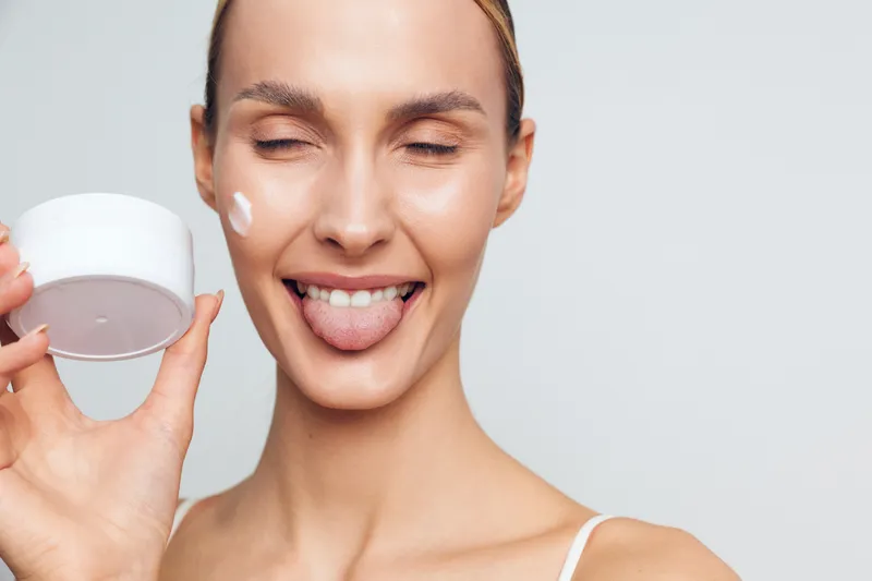A smiling young woman applies white cream on her cheek while holding a jar of skincare product. She sticks out her tongue playfully against a plain light background.