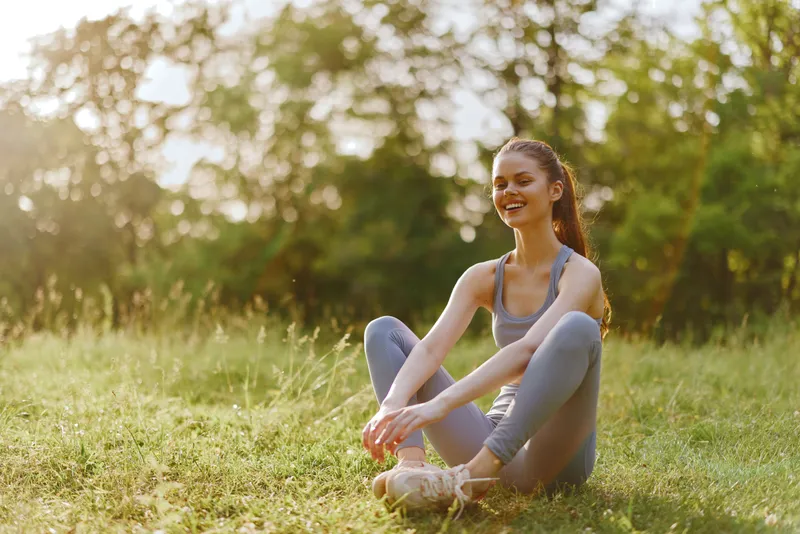 Happy young woman in activewear sitting on grass, enjoying sunlight in a natural park, representing vitality, fitness, and outdoor lifestyle