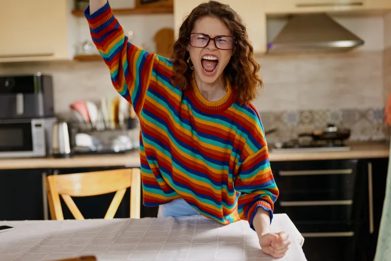Young woman with curly hair and glasses celebrates at home in a bright kitchen, wearing a rainbow striped sweater, smiling with energy and joy while standing beside a wooden table