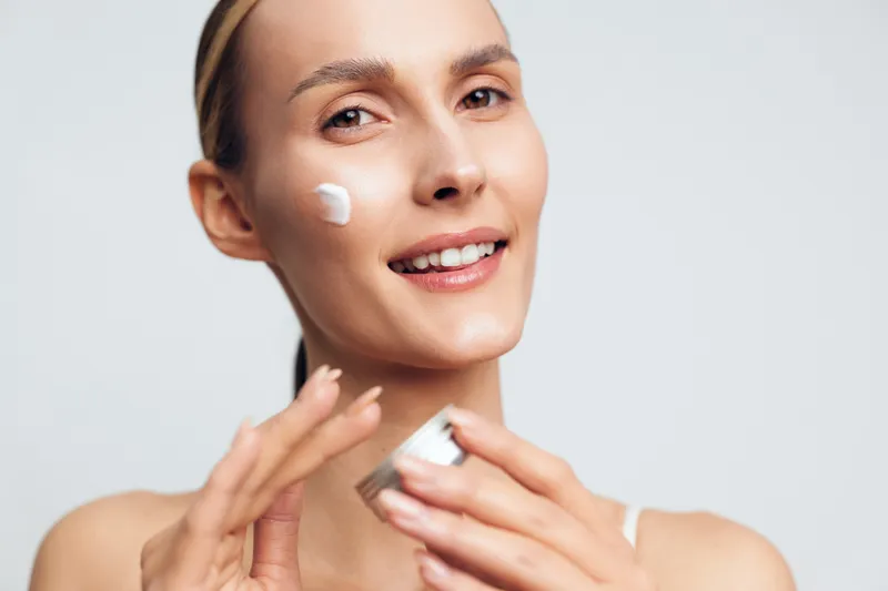 Bright portrait of a smiling woman applying moisturizer to her cheek, clean skincare moment, soft lighting, healthy glow, natural expression, minimal makeup, fresh complexion