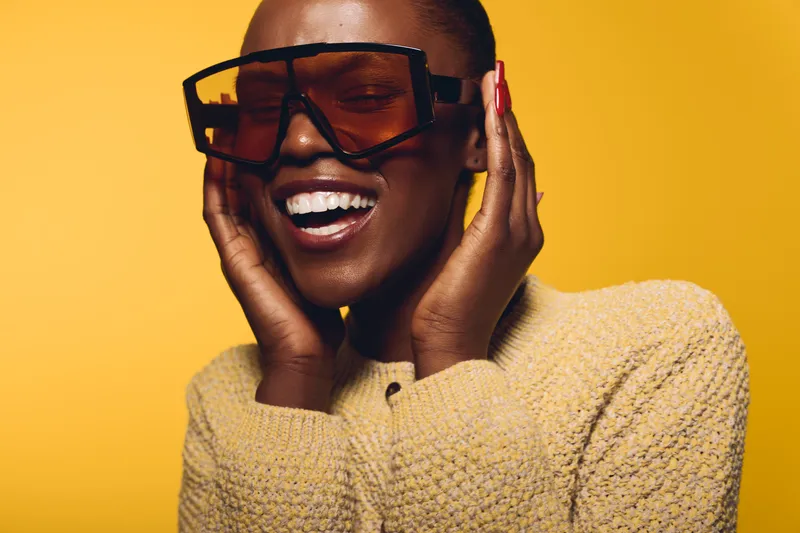 Happy dark skinned African American woman wearing oversized sunglasses and a beige sweater smiles with closed eyes isolated on yellow background. Concept of joy and fashion.