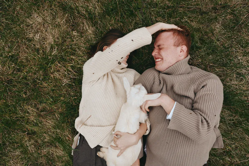 Couple enjoys a cozy lifestyle moment outdoors on grass, cuddling a small animal, smiling as they share affection with a gentle goat kid, warm sweaters creating a relaxed, intimate scene against a