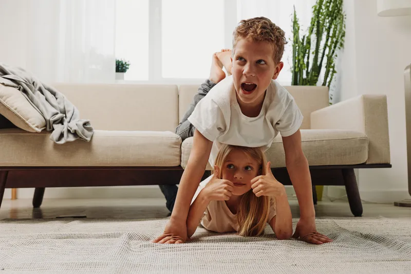 Playful children having fun together at home, boy balancing on girl lying on floor, bright living room with natural light and casual cozy atmosphere.