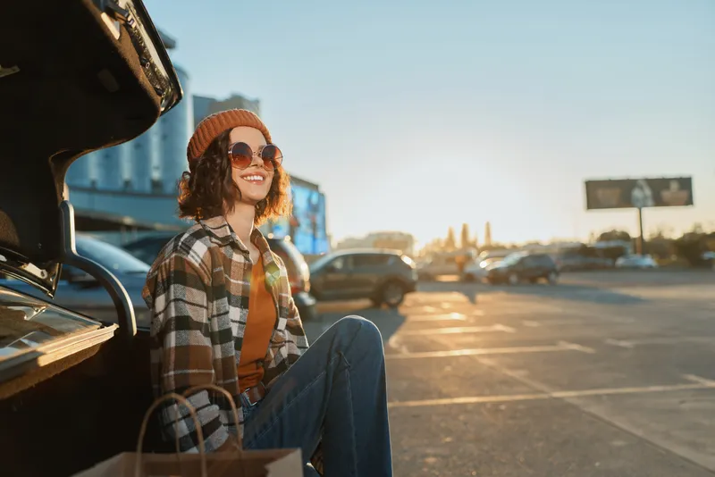 woman smile car parking shopping sunset sunglasses beanie sitting in trunk with shopping bags, candid lifestyle and authenticity captured in golden hour glow, mindful living and emotional storytelling