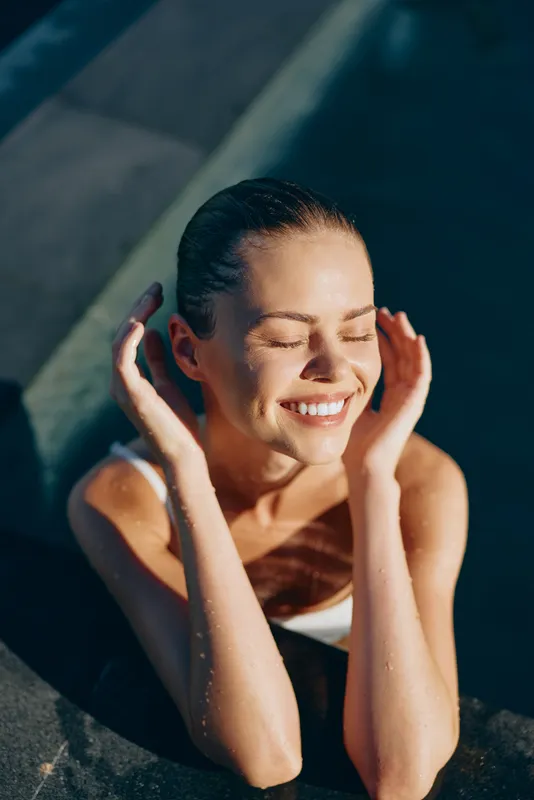Smiling woman in a swimsuit rests at the pool edge with eyes closed, enjoying warm sunlight. Concept of wellness, relaxation, and leisure moments.
