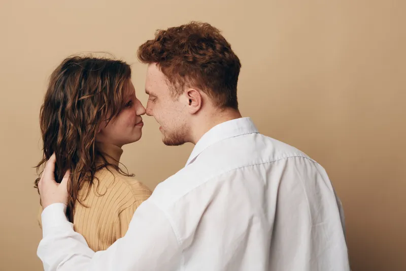 Young couple embraces tenderly facing each other against beige background. Intimate moment shows affection, love, and connection between man and woman in casual clothing.